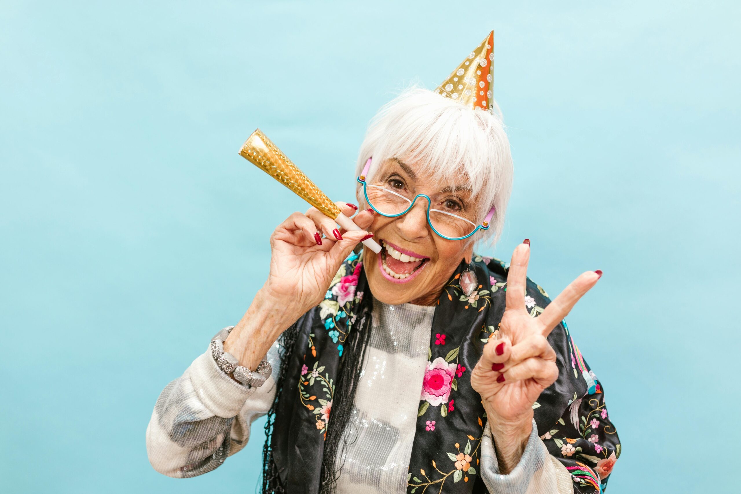 Senior woman in party hat joyfully celebrating, making peace sign on a light blue background.
